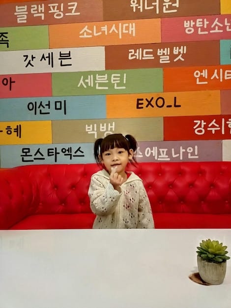 A split-screen photograph. On the left, a vibrant and modern Korean language school class in Seoul, South Korea. On the right, an image of a young Brasilian woman immersed in studying Korean with books and online resources.