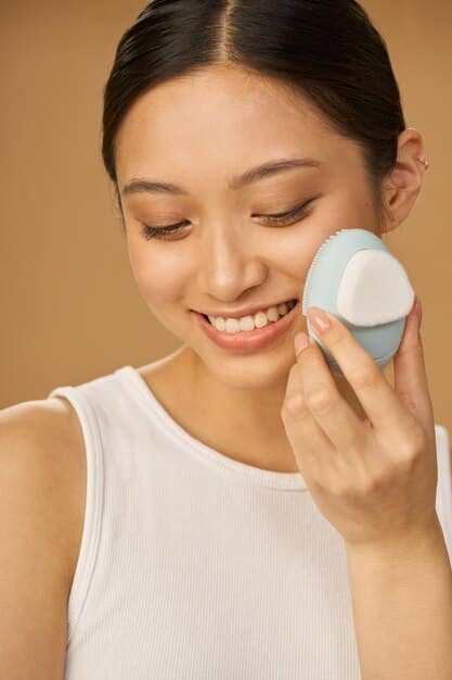 A Korean woman applying sunscreen on her face with a sponge applicator, her other hand holding the sunscreen bottle. She is smiling and standing in front of a window with natural light. The focus is on her face and the application of the product.