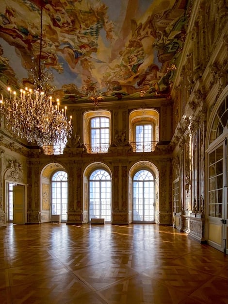 A wide shot of a grand ballroom inside a European palace during the 18th century. The ballroom is brightly lit by numerous crystal chandeliers, with ornate decorations and gilded details adorning the walls and ceiling. Men in formal attire and women in elegant, colorful gowns are dancing gracefully. Musicians play classical music on a raised platform at one end of the hall.